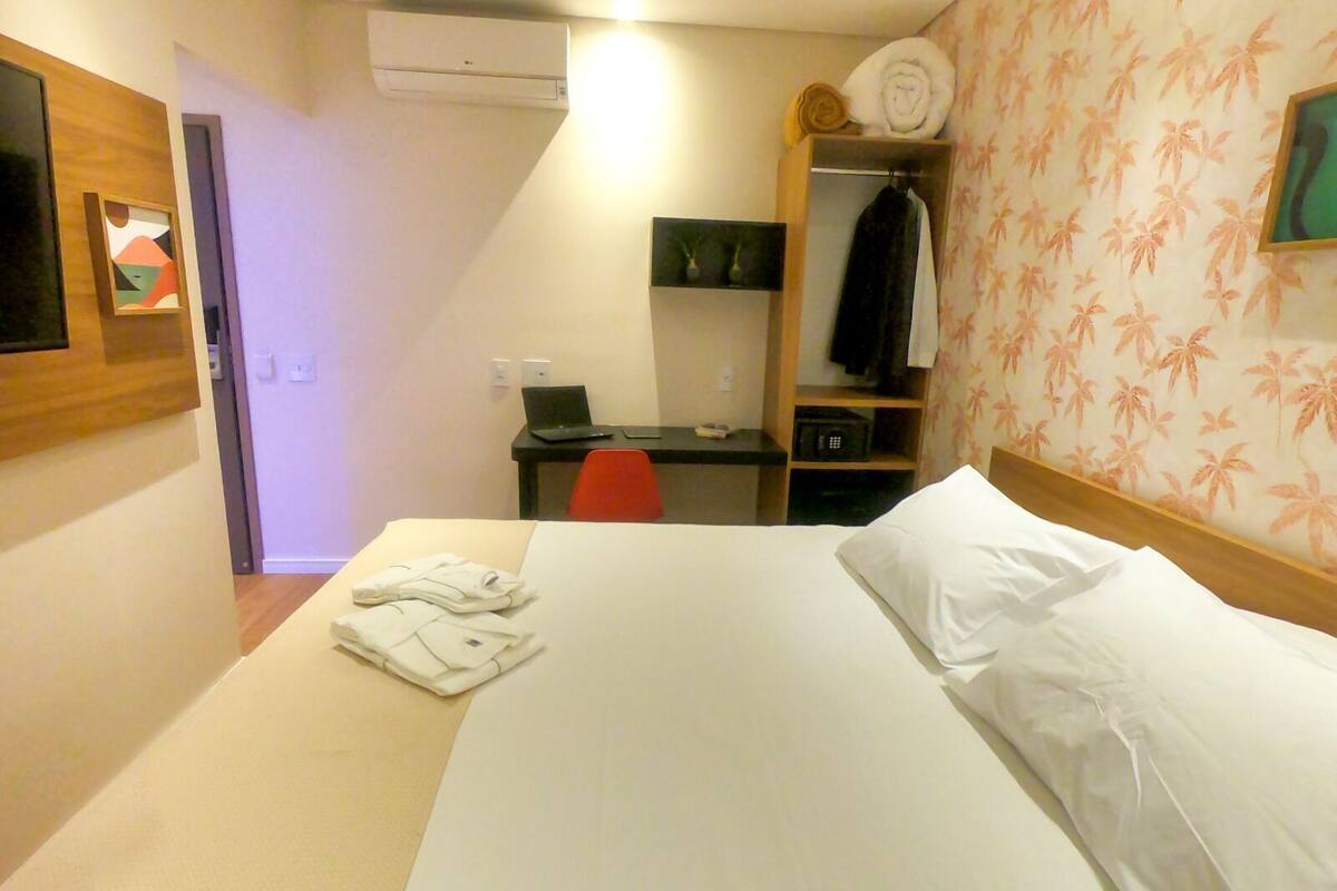 A hotel room with a bed, a desk with a laptop, a chair, a wardrobe, and a wall-mounted air conditioner.