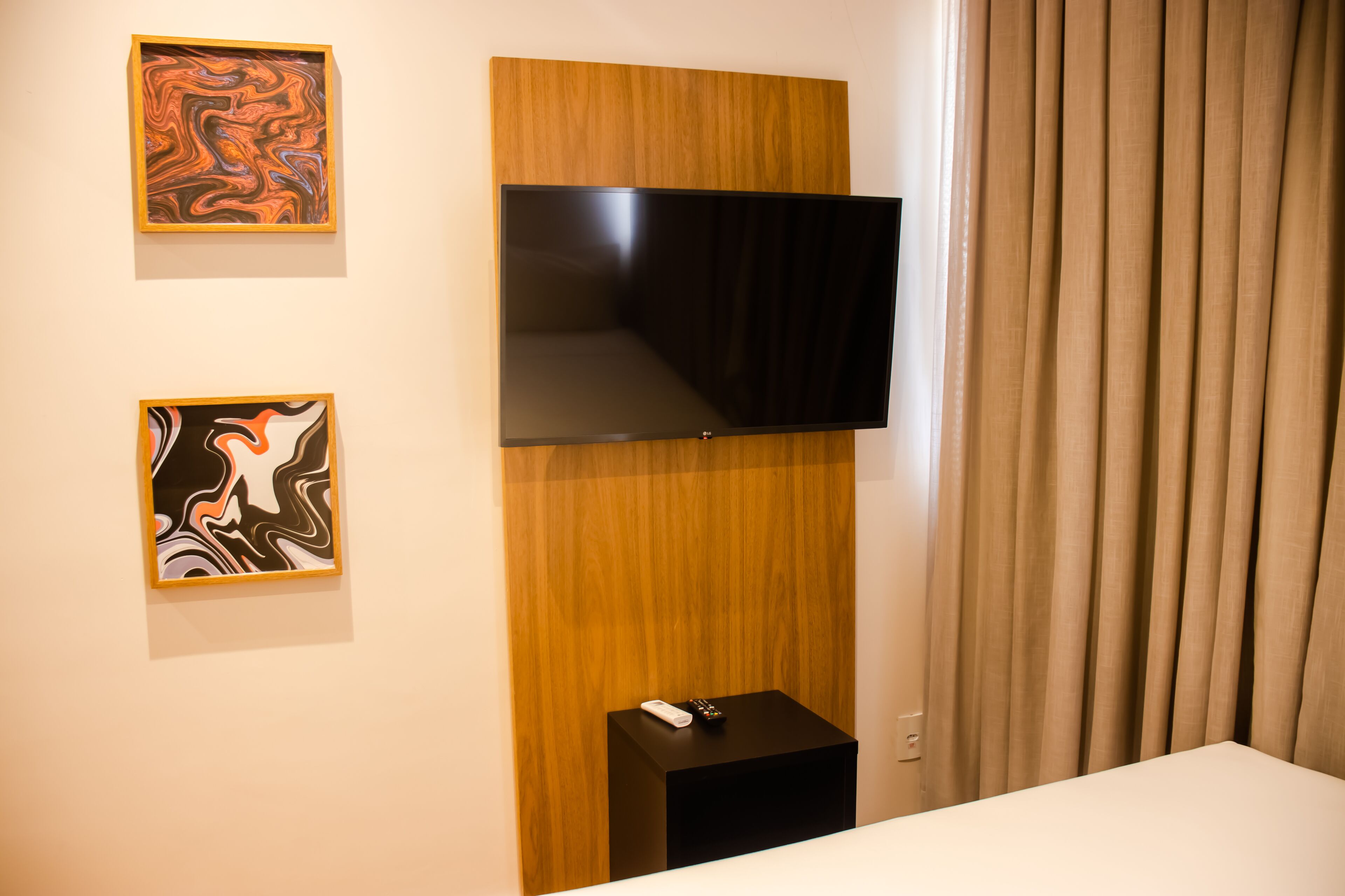 A hotel room with a flat-screen TV mounted on a wooden panel, a small black side table with a remote control, and two abstract paintings on the wall.