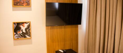 A hotel room with a flat-screen TV mounted on a wooden panel, a small black side table with a remote control, and two abstract paintings on the wall.