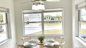 Comedor con una mesa redonda dispuesta para cuatro personas, rodeada de sillas blancas con asientos acolchados. Un ventilador de techo se encuentra sobre la mesa, y hay ventanas amplias que ofrecen vistas al exterior.