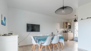 A modern dining area with a TV mounted on the wall, a dining table with chairs, and a kitchen area visible through an open door. - La Marine - Jardin - Golf (Pornic)