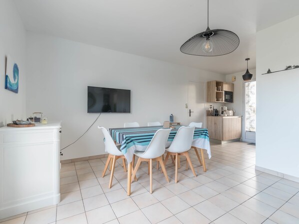 A modern dining area with a TV mounted on the wall, a dining table with chairs, and a kitchen area visible through an open door. - La Marine - Jardin - Golf (Pornic)