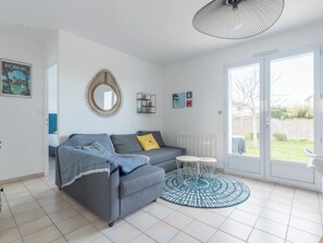 Un salon moderne avec un canapé gris, une table basse ronde, un tapis à motifs, un miroir décoratif et une grande fenêtre donnant sur l’extérieur.
