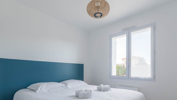 A modern bedroom with a large bed, white bedding, a blue accent wall, a window with a view, and a woven ceiling light fixture. - La Marine - Jardin - Golf (Pornic)