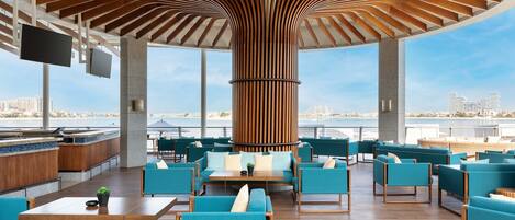 A modern outdoor seating area with wooden beams, blue upholstered furniture, and a view of the city and water.