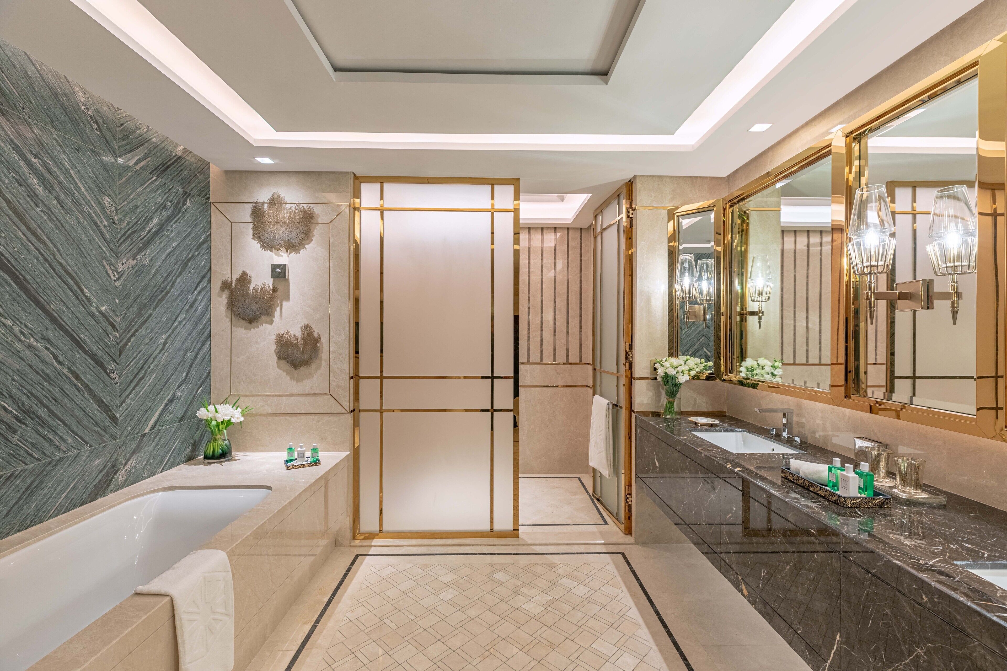 A modern bathroom with a large bathtub, two sinks, a mirror, and decorative elements like flowers and a vase.