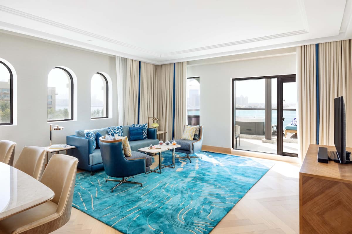 A modern hotel room with a large balcony, a blue sofa set, a dining area, and a view of the city.