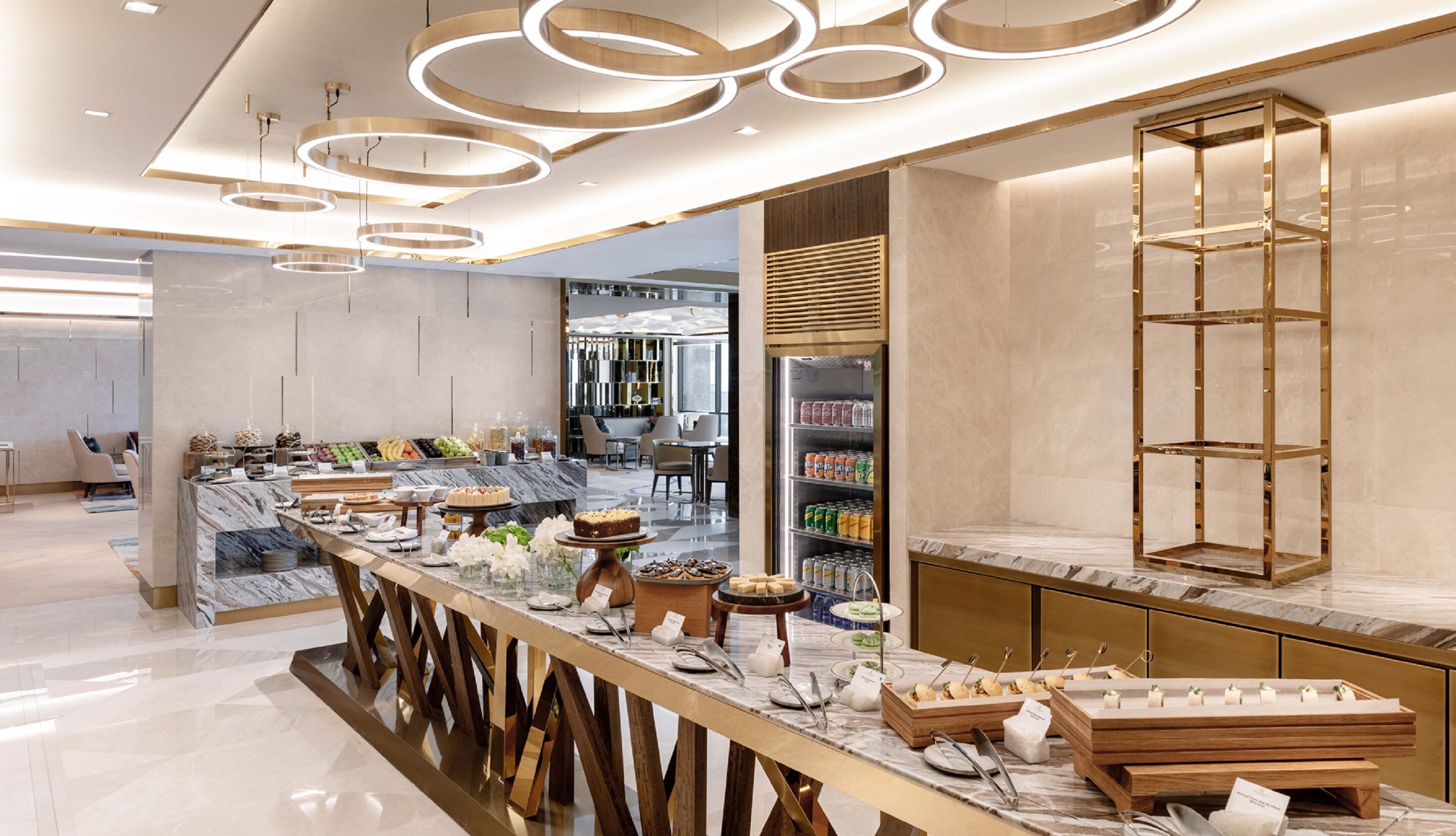 A modern hotel lobby with a marble reception desk, a buffet area with various food items, and a seating area with chairs and tables.