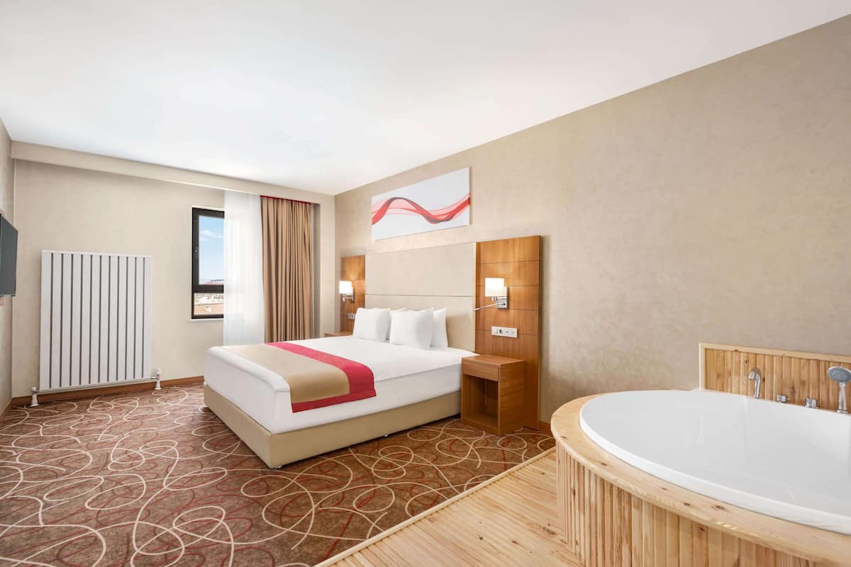 Een hotelkamer met een groot bed, een houten hoofdeinde, een wandtelevisie, een raam met gordijnen, een badkamer met een bad en een houten vloer.