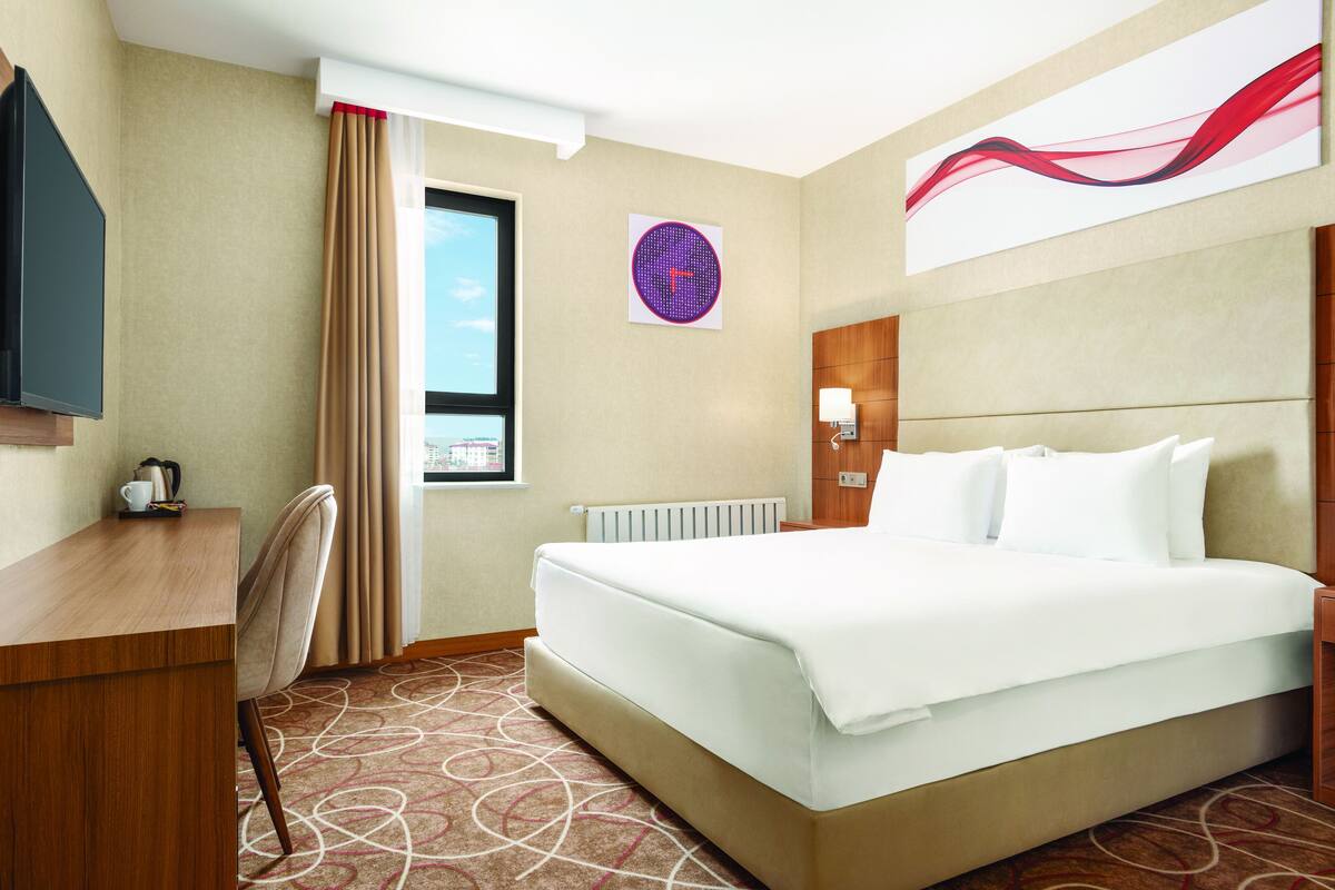 Hotelkamer met een bed, bureau, stoel, raam, radiator en een wandklok.