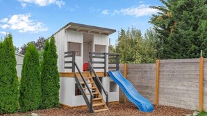 Una casita de juegos de madera con un tobogán, rodeada por una valla de madera y árboles altos.