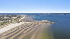 Ein Luftbild einer Küstenregion mit einer langen Pier, die sich ins Wasser erstreckt, einem Sandstrand mit deutlichen Sandmustern und einer Stadt mit Gebäuden im Hintergrund.