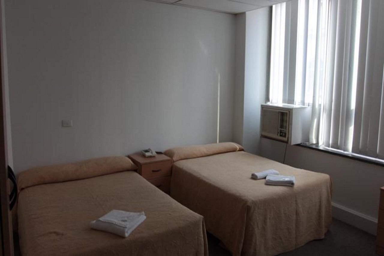 Habitación de hotel con dos camas individuales, una mesita de noche con teléfono, una ventana con cortinas y un equipo de aire acondicionado instalado en la pared.