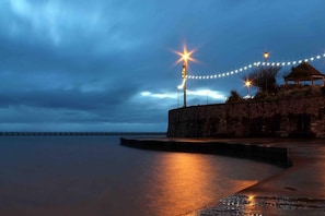 A waterfront promenade at dusk with stone walls and string lights.