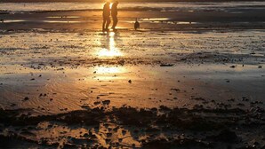 Rantamaisema auringonlaskun aikaan, jossa kaksi ihmistä seisoo ja lapsi kävelee taustalla.