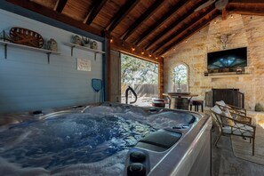 Ein Whirlpool in einem rustikalen Raum mit Steinwänden, Holzbalken, einem Kamin aus Stein und Regalen mit Dekorationsgegenständen.