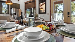 Una mesa de comedor servida con platos, tazones y vasos blancos, una botella de vino y una fuente de frutas.