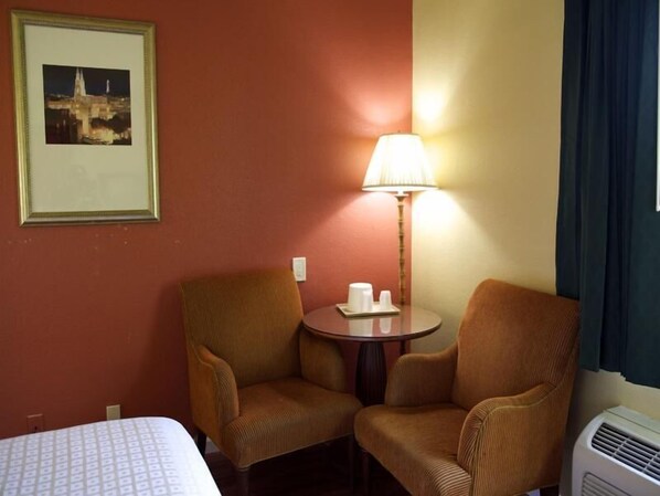 A hotel room with two armchairs, a small round table with a lamp, a bed, and a wall-mounted air conditioner. - Sundial Lodge (Redding)