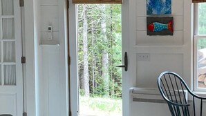 A room with a door leading to a forest view, a thermostat, and a chair. - Charming and cozy apartment nestled in the woods. (Mount Desert)