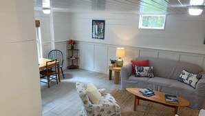 A cozy living room with a striped sofa, a small table, and a chair. There is a window with a view of trees and a painting on the wall. - Charming and cozy apartment nestled in the woods. (Mount Desert)