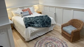 A bedroom with a bed, a bedside table, a lamp, a radiator, a wicker chair, and a patterned rug. - Charming and cozy apartment nestled in the woods. (Mount Desert)