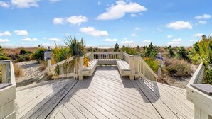 Ein hölzerner Steg führt zu einem Strand mit blauem Himmel und spärlicher Vegetation.