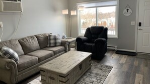 A living room with a gray sofa, a black recliner, a wooden coffee table, and a large window. - Adventure Acres- Suite B2 (Holly)