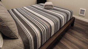 Quarto com cama, televisão, janela com persianas, mesa de cabeceira e um sistema de ventilação na parede.