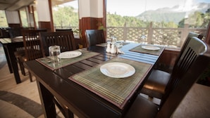 A dining table set with plates, glasses, and salt shakers, with a view of trees and mountains in the background. - Aamod @ Bhimtal (Nainital)