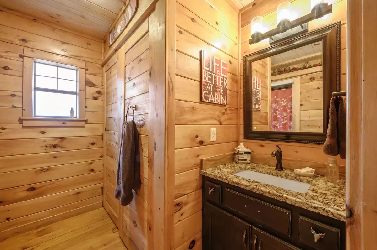 A wooden cabin bathroom with a window, a sink with a mirror, and a towel hanging on the wall.