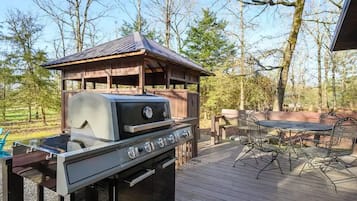 Ein Grill auf einer Holzterrasse, mit Tisch und Stühlen, umgeben von Bäumen.