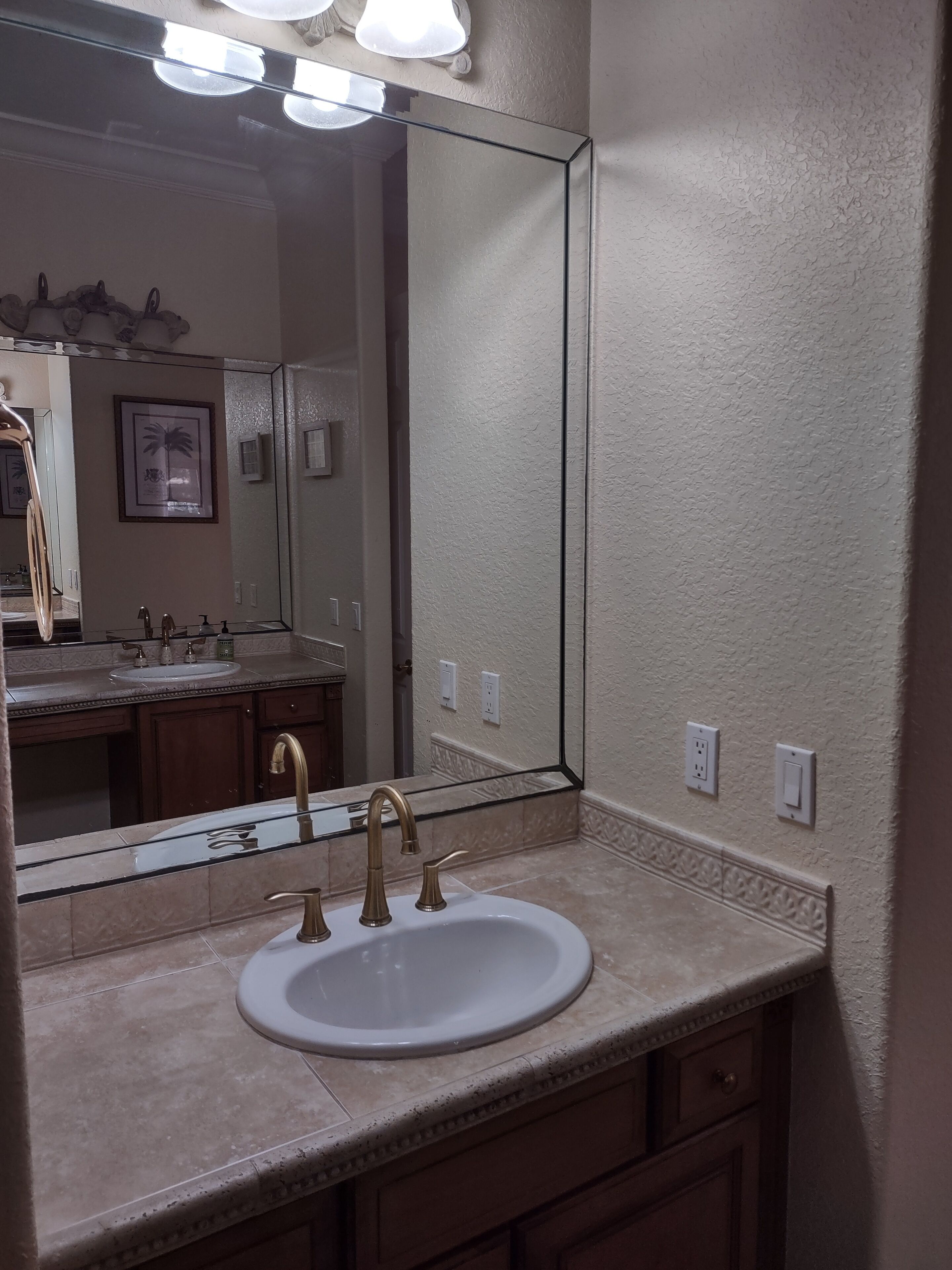 Un bagno con uno specchio grande, un lavandino bianco con rubinetteria dorata e un piano di lavoro con bordo decorativo.