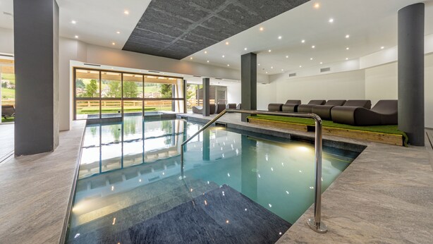 Un espace piscine intérieur moderne, doté d’une paroi en verre, d’un coin salon et bénéficiant d’une lumière naturelle grâce à de grandes fenêtres.
