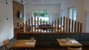 Un espace de restauration moderne avec des tables et des chaises en bois, un banc vert rembourré et une vue sur l’extérieur grâce à de grandes fenêtres.
