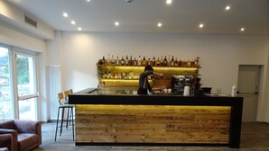 Un espace bar moderne avec un comptoir en bois, une machine à café et diverses bouteilles. On peut voir au premier plan deux chaises en bois et un canapé.