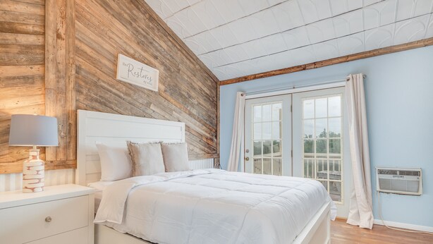 Ein Schlafzimmer mit einer Holzverkleidung, einem weißen Bett, einem Nachttisch, einer Lampe, einem Fenster mit Vorhängen und Blick ins Freie.