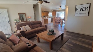 Un salón espacioso con un sofá de cuero marrón, una mesa de centro de madera y una zona de comedor con sillas de madera. Se ve una cocina al fondo.