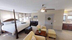 Ein Schlafzimmer mit einem Himmelbett, einem Deckenventilator, einem Schreibtisch mit Stuhl, einem kleinen Tisch mit Pflanze und einer Tür mit einem Kranz.