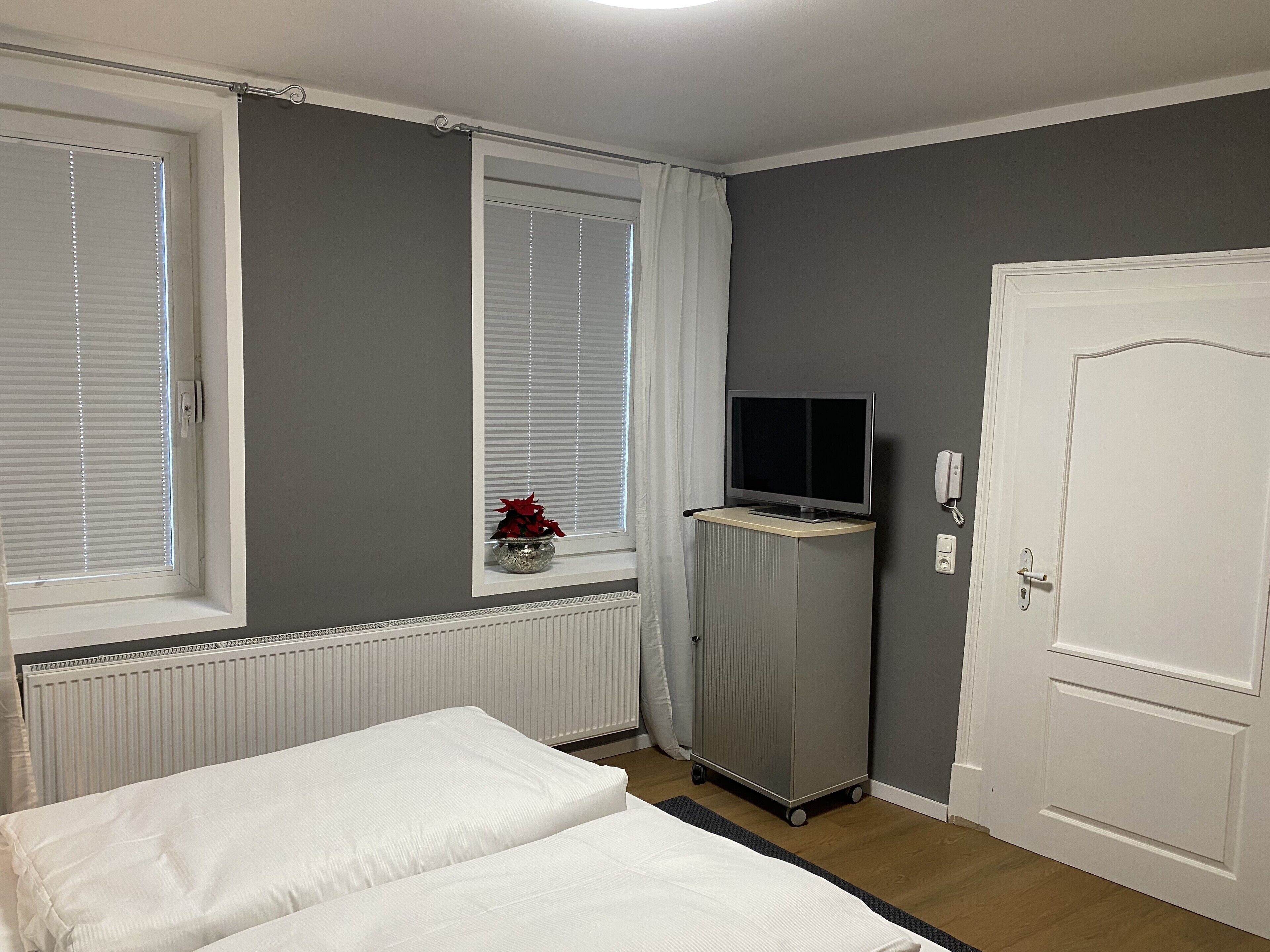 A hotel room with a bed, a TV on a stand, a door, and a window with blinds.