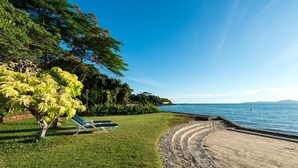 A beach with a stone pathway leading to the water, a grassy area with a tree and two lounge chairs, and a clear view of the sea and distant land. - RCG Suites Pattaya (Pattaya)