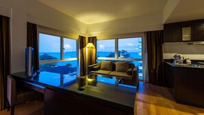 A modern hotel room with a glass dining table, a sofa, and a large window offering a view of the ocean and cityscape. - RCG Suites Pattaya (Pattaya)