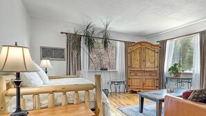 A cozy bedroom with a wooden bed, bedside lamps, a wooden nightstand, a wooden bench, a wooden table, a wooden cabinet, a potted plant, and a window with curtains. - Rocky Mountain Inn (Paonia)