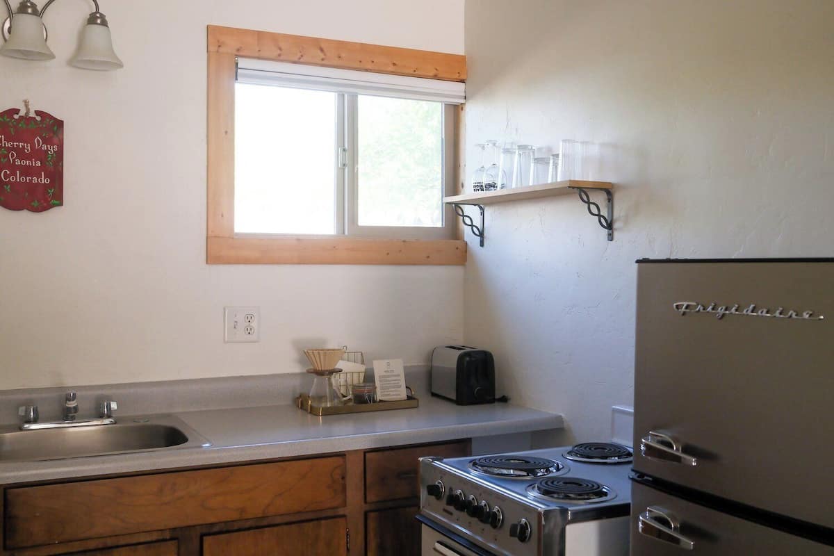 A kitchen with a stove, sink, and a window above the counter. There are two light fixtures on the wall and a shelf with glassware.