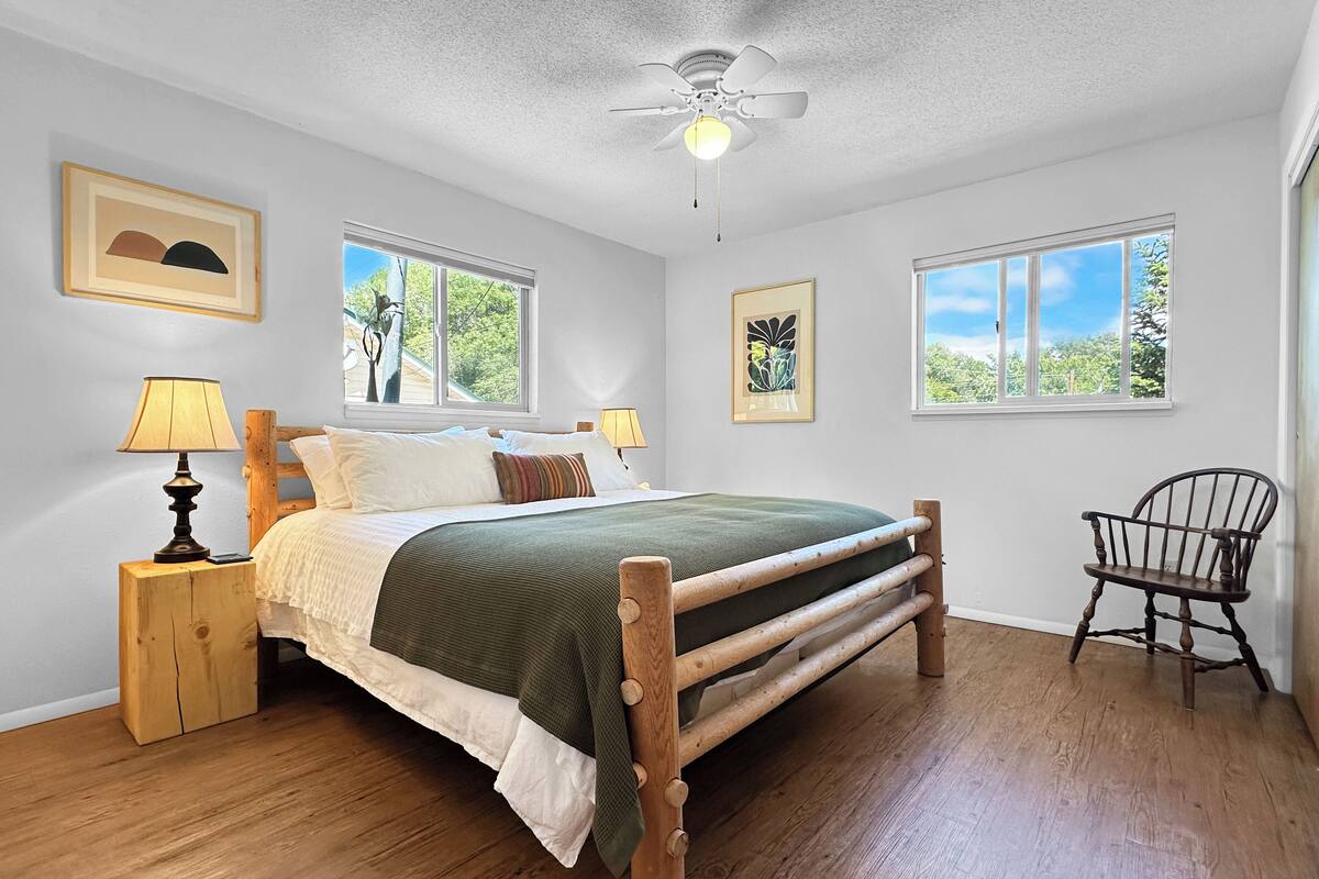 A bedroom with a wooden bed, two bedside tables with lamps, a chair, and two windows with views of greenery.