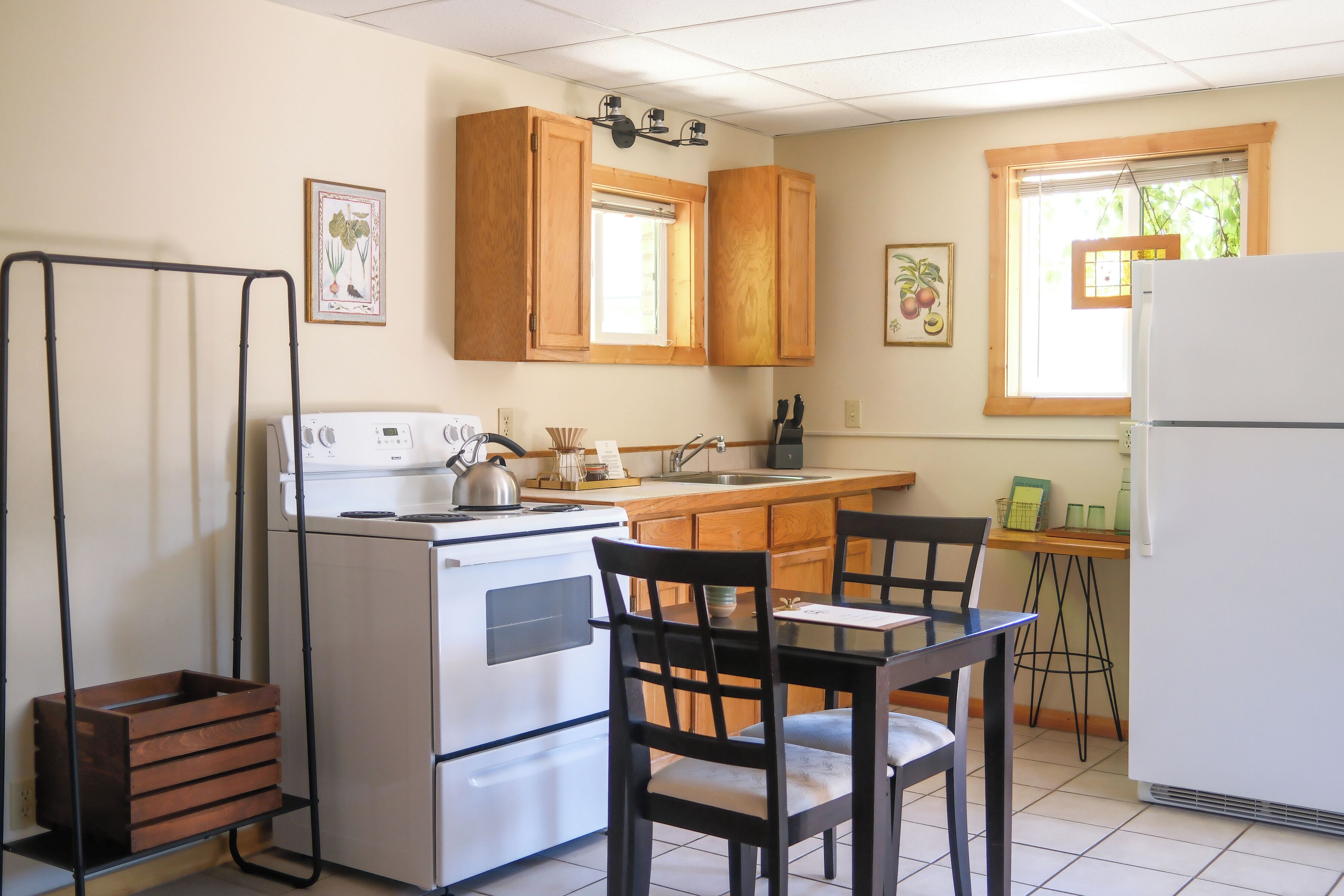 A compact kitchen with a stove, sink, and refrigerator. There are wooden cabinets, a window with blinds, and a small dining area with chairs.