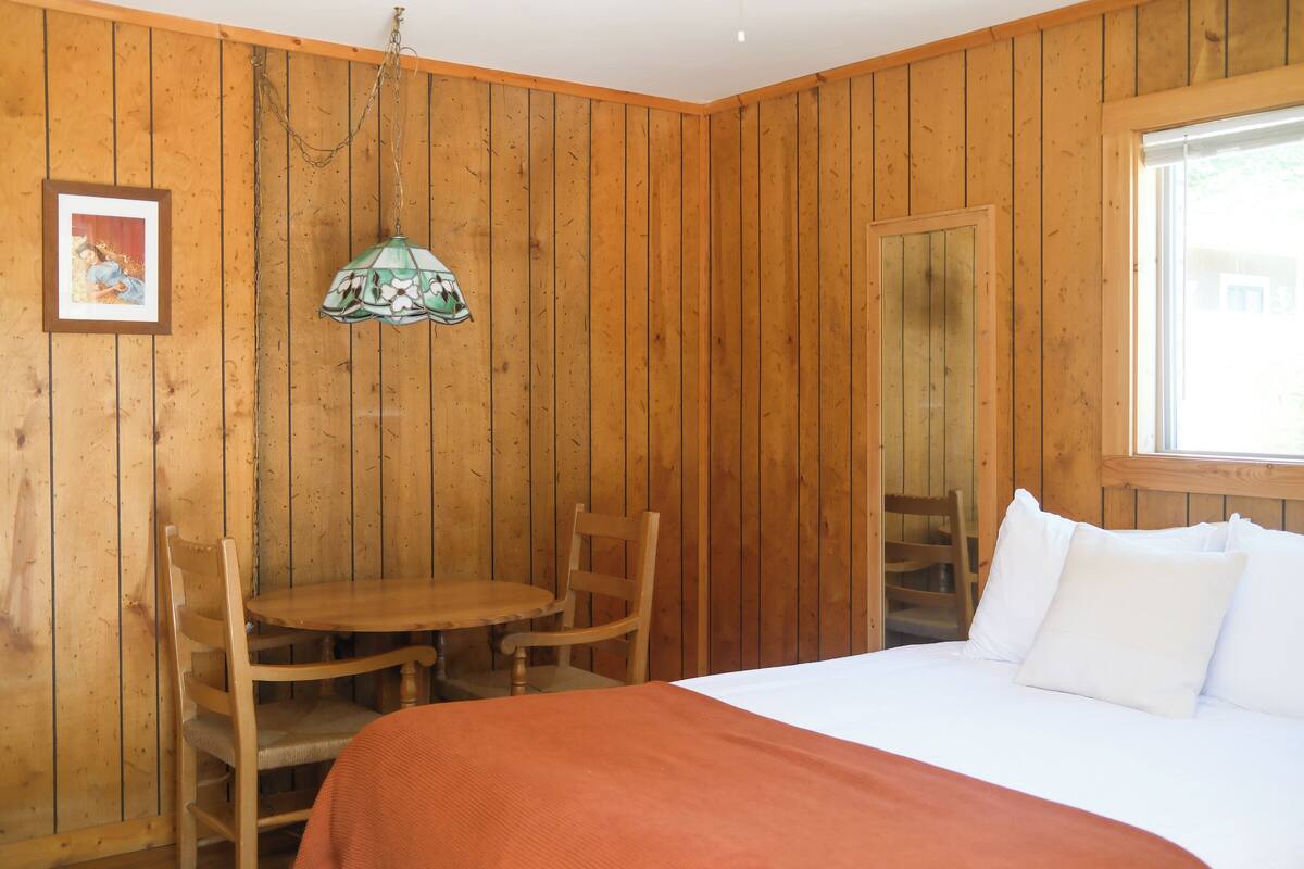 A wooden paneled room with a bed, a table, and two chairs.