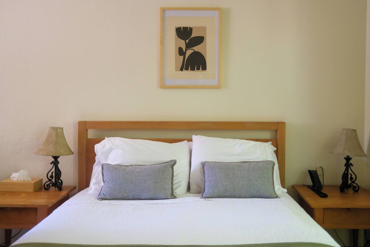 A neatly made bed with white linens and two grey pillows, flanked by wooden nightstands with lamps and a framed artwork above.