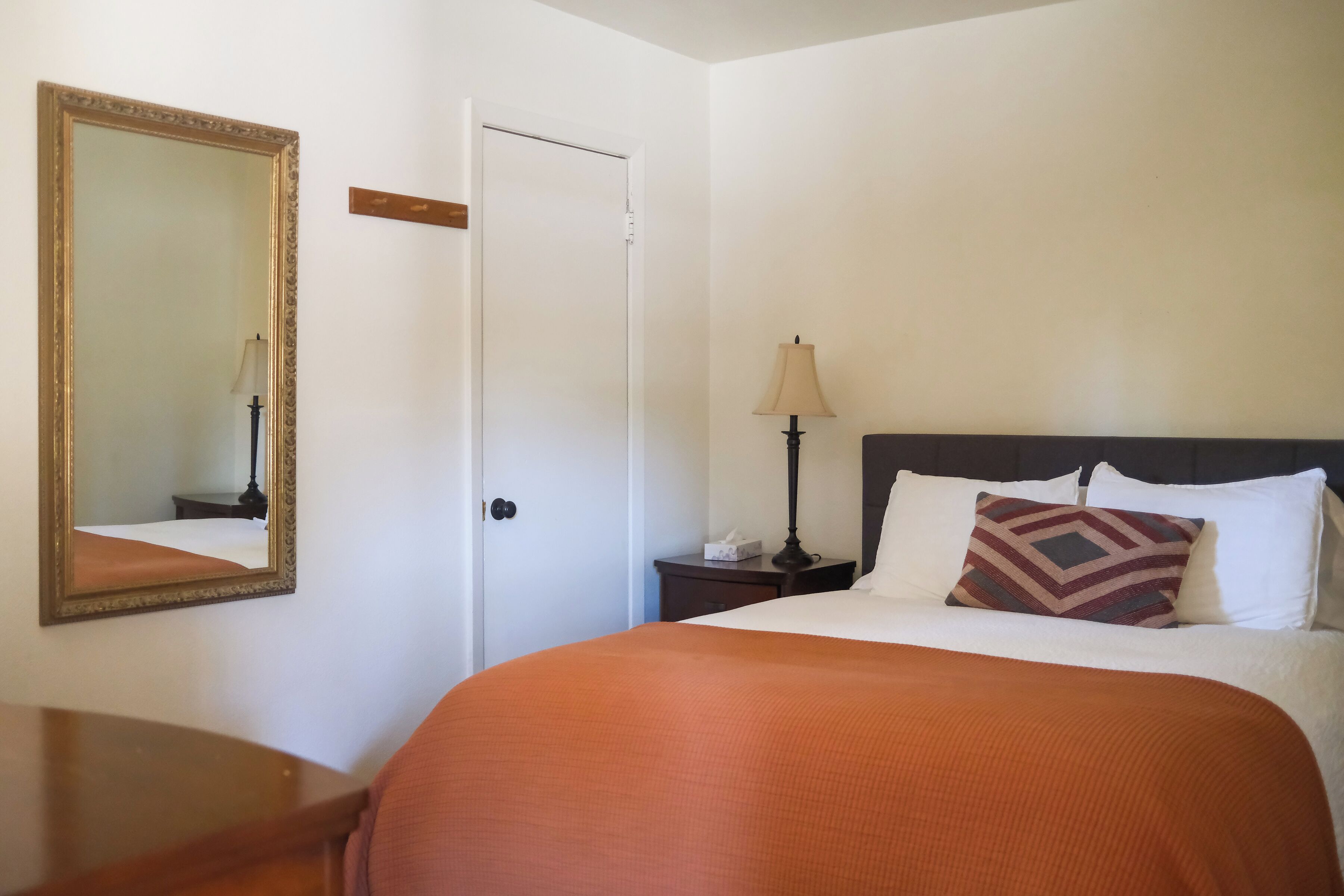 A hotel room with a bed, a nightstand, a lamp, a mirror, and a door.