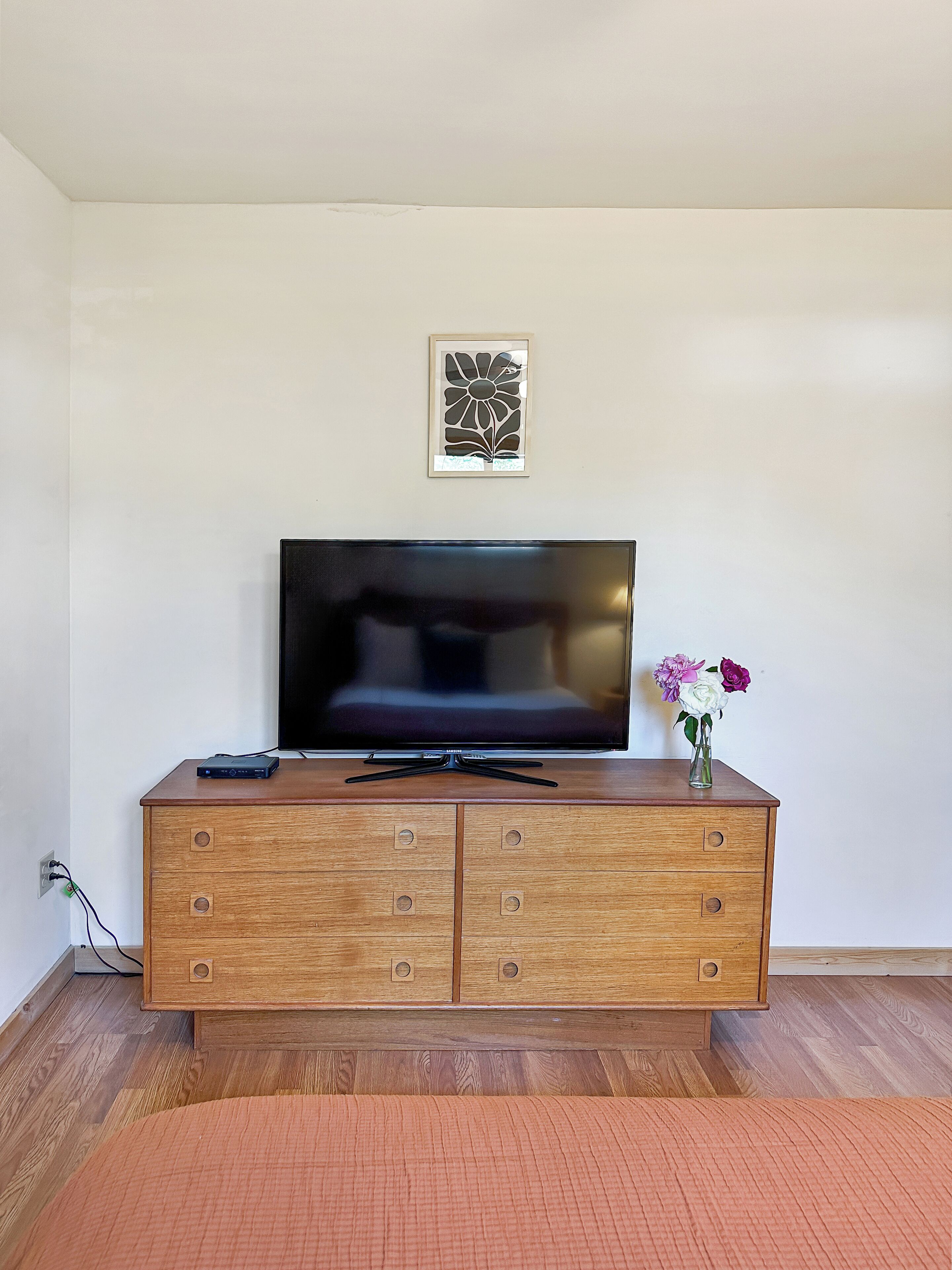 Un mueble bajo de madera para televisor con un televisor de pantalla plana, una obra de arte enmarcada encima y un florero con flores a la derecha.