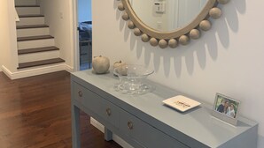 A hallway with a staircase, a round mirror with a wooden frame, a light blue console table with a clear glass bowl, and a framed photo. - In Sag Harbor Village. Walk to town, and less than 1/2 mile to Havens Beach. (Sag Harbor)